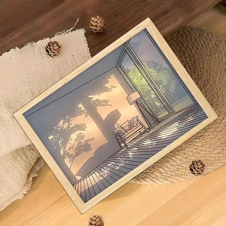 Framed artwork of a house exterior on a wooden surface with pine cones.