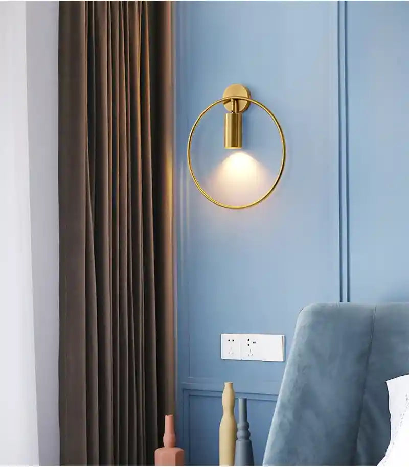 Gold circular wall light fixture on a blue paneled wall with a curtain and chair in the background.