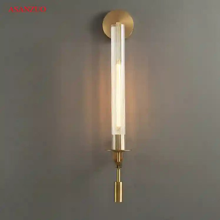 Gold wall sconce with clear glass shade on a gray wall, featuring the ASANZIO brand.