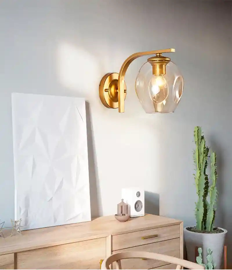 Gold wall sconce with glass shade in a room setting with a plant and furniture.