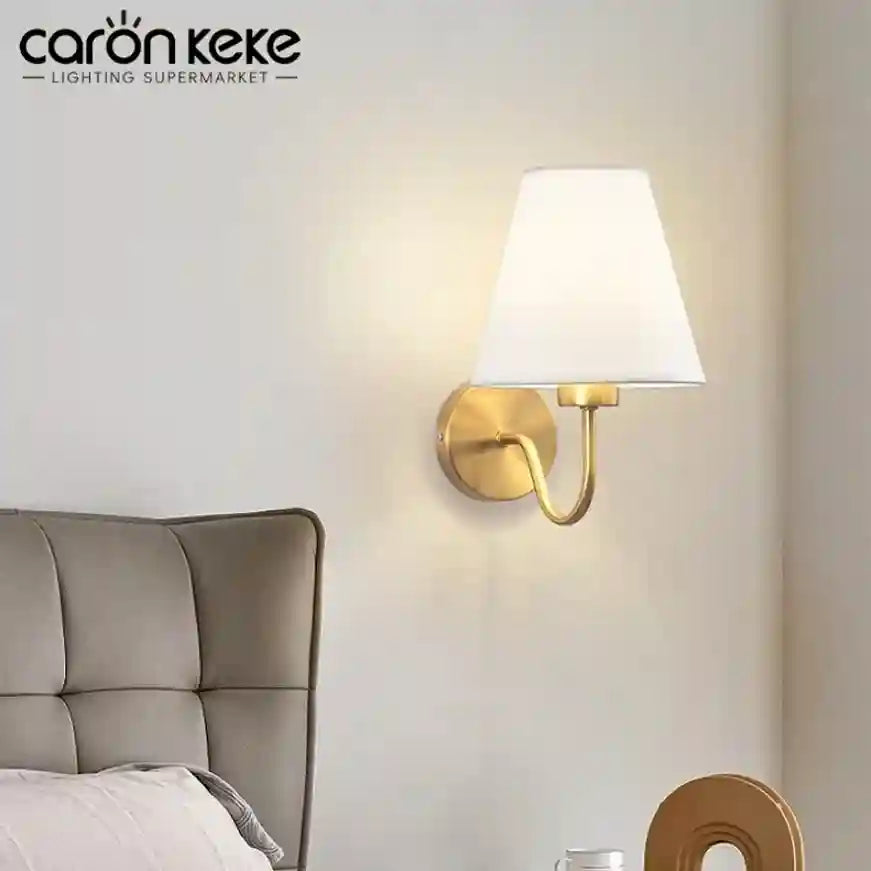 Wall-mounted lamp with a white shade and gold fixture on a wall next to a bed, branded 'caron keke'.