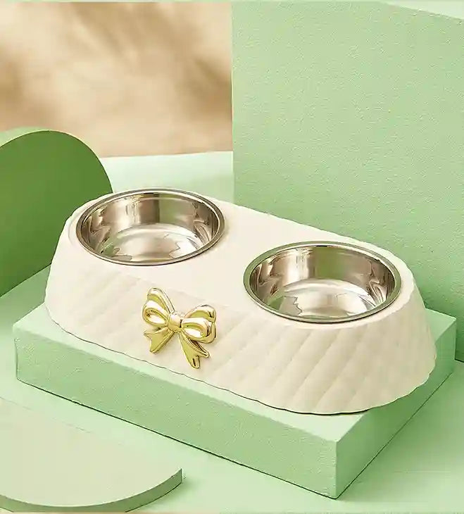 White pet bowl with two silver bowls and a gold bow on a green platform.